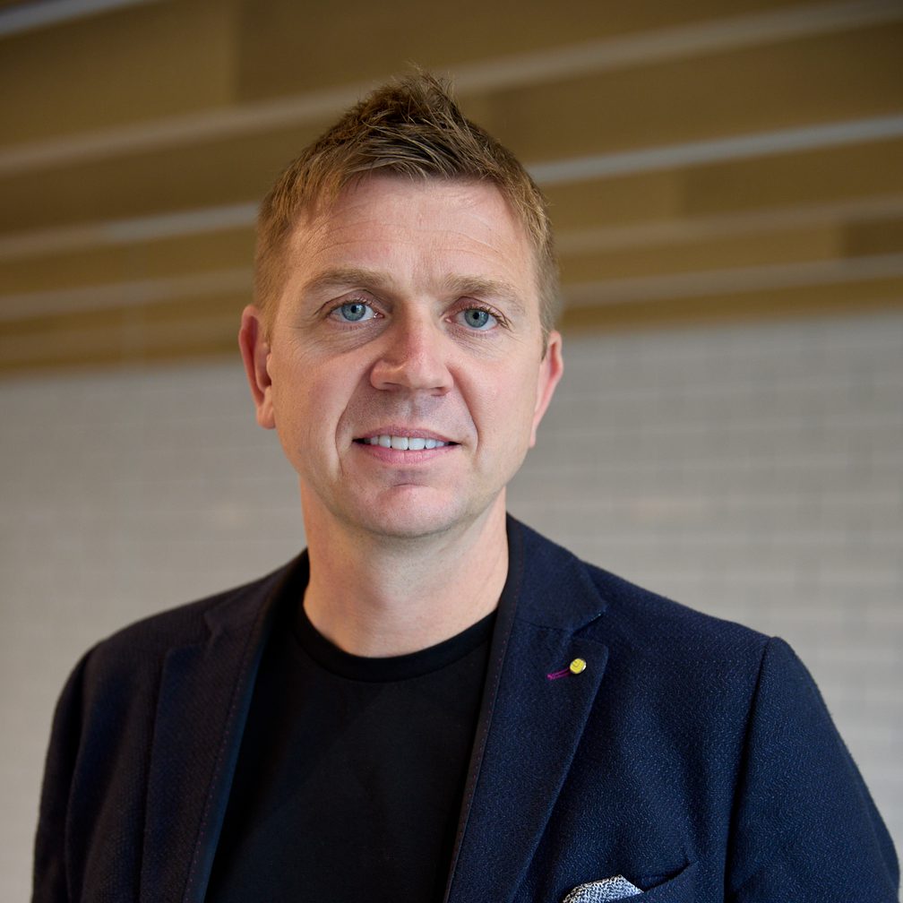 Person with short light brown hair and blue eyes, wearing a navy blazer, black top and pocket square, indoors with a light tiled wall backdrop.