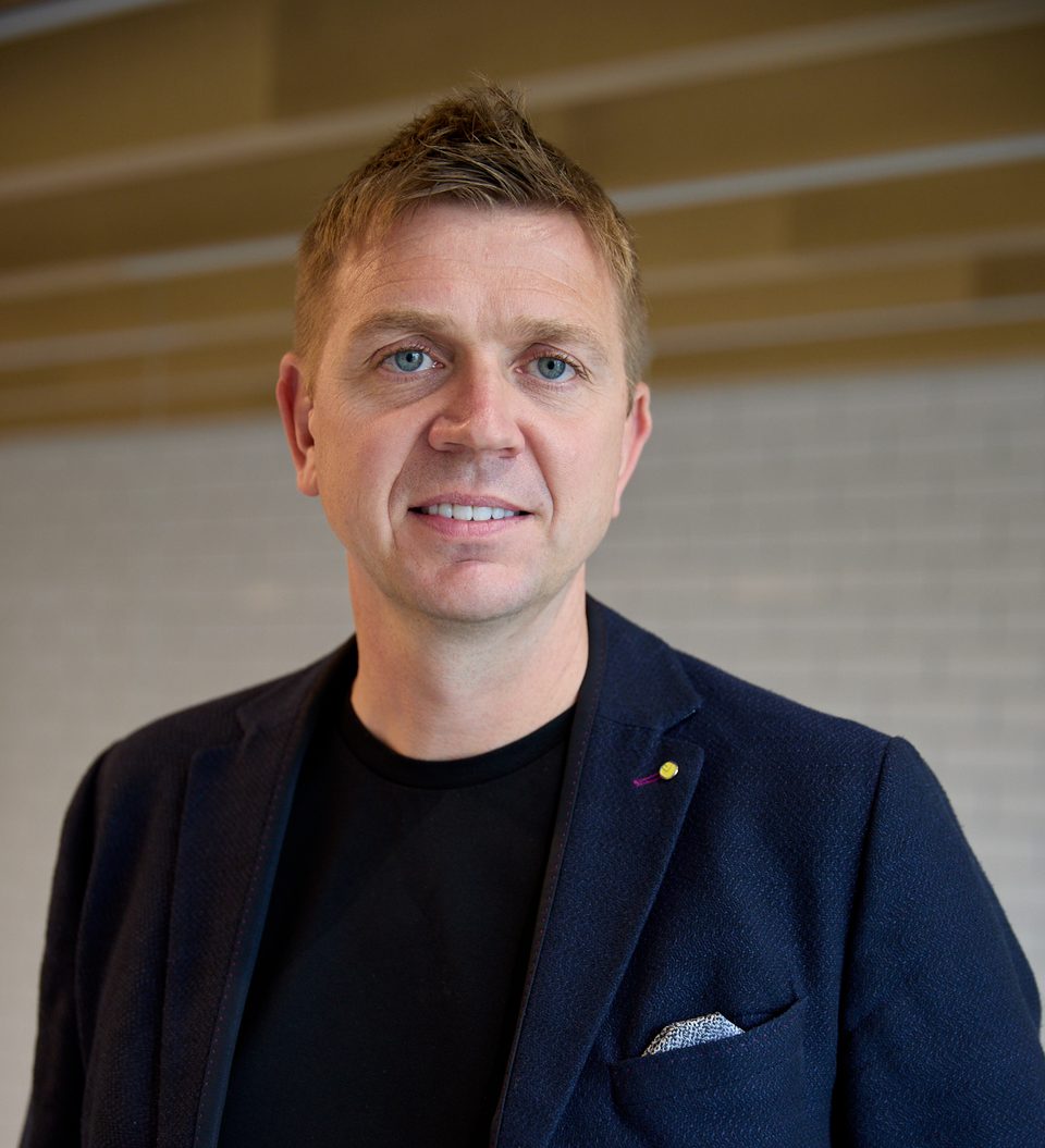 Person with short light brown hair and blue eyes, wearing a navy blazer, black top and pocket square, indoors with a light tiled wall backdrop.