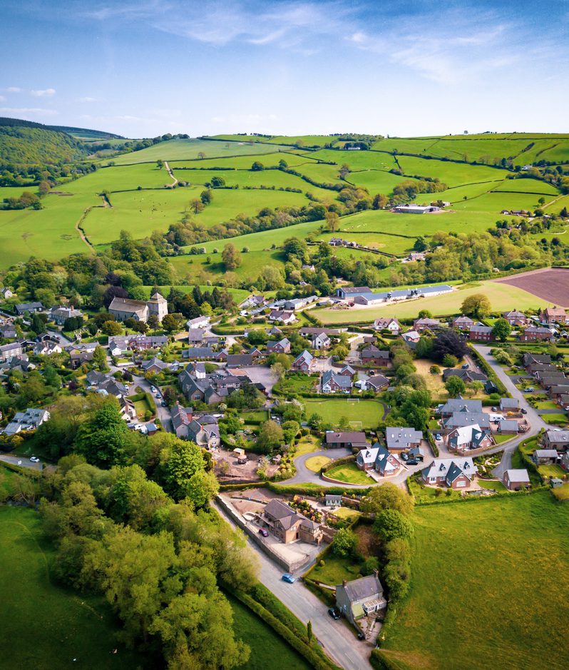 Aerial view of a small village nestled in rolling green countryside, with hedges, fields, winding lanes and farm buildings under a bright blue sky.